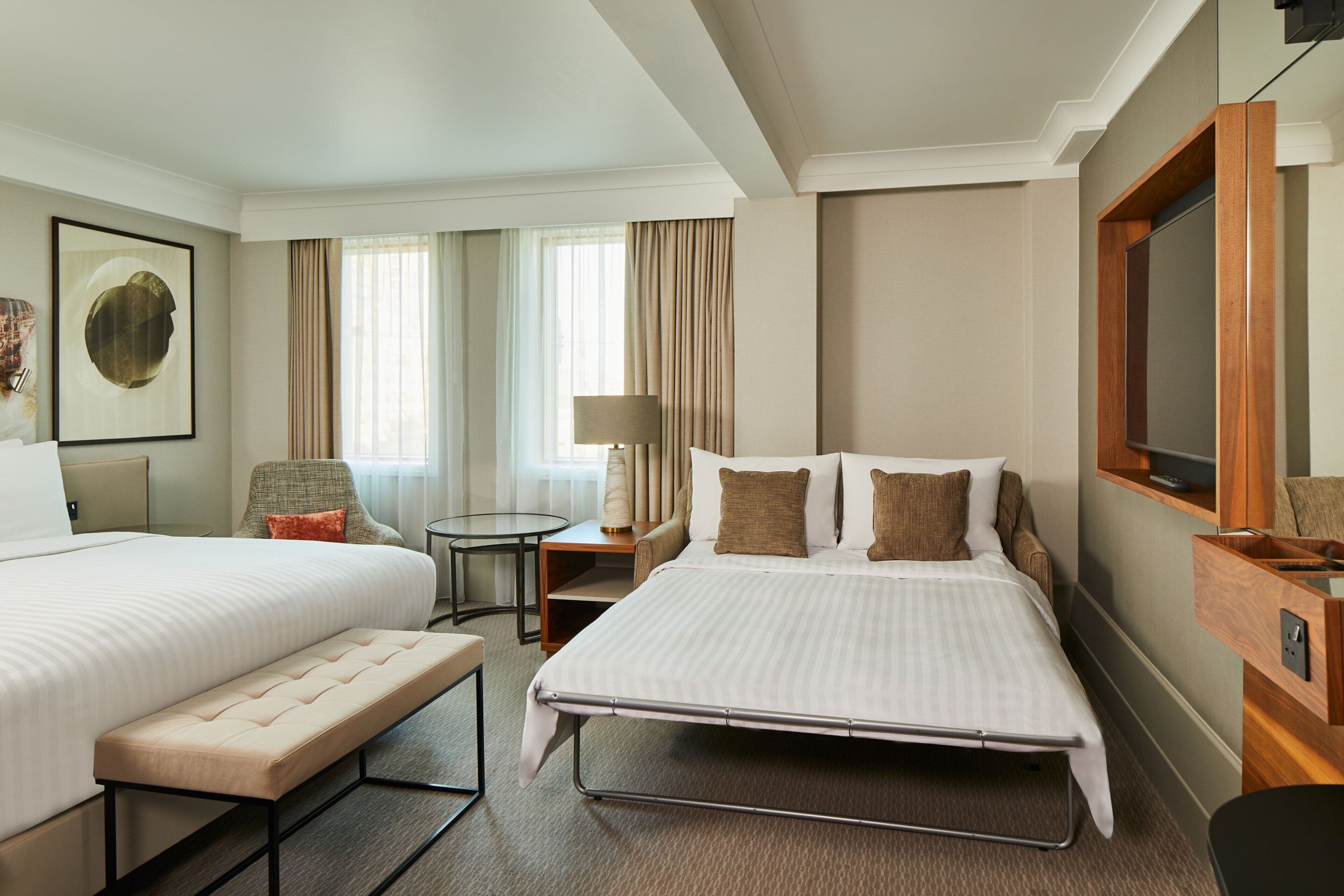 A hotel room with two beds, a bench, a desk, a chair, a lamp, and a TV.