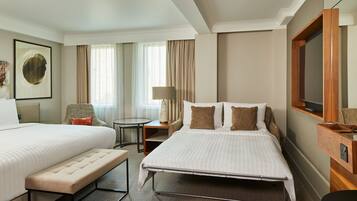 A hotel room with two beds, a bench, a desk, a chair, a lamp, and a TV.