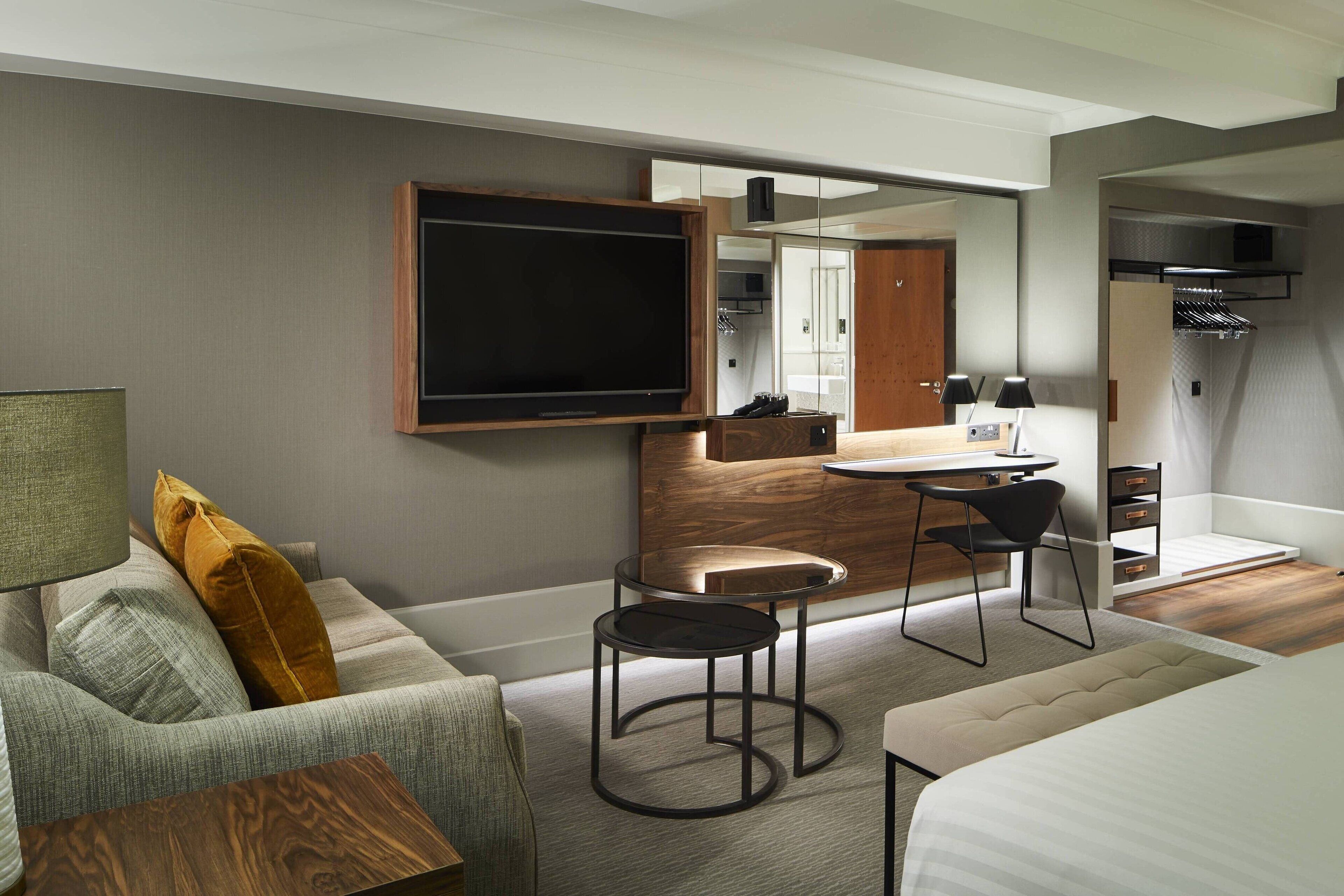 A modern hotel room with a large flat-screen TV mounted on the wall, a sofa with cushions, a small round table, a chair, a bed, and a desk with a chair.