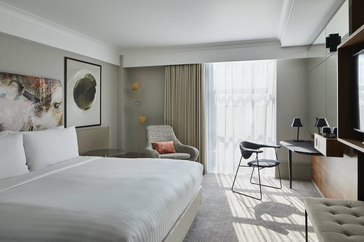 A hotel room with a large bed, a desk, a chair, and a sofa. There is a window with curtains, a painting on the wall, and a lamp.