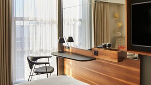 A hotel room with a large window, a desk with a chair, a bed, and a TV mounted on the wall. - London Marriott Hotel Kensington (London)