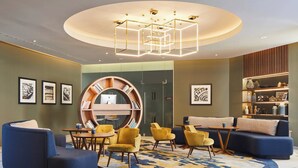 A modern lounge area with a circular seating arrangement, a central bookshelf, and a unique geometric ceiling light fixture. - London Marriott Hotel Kensington (London)
