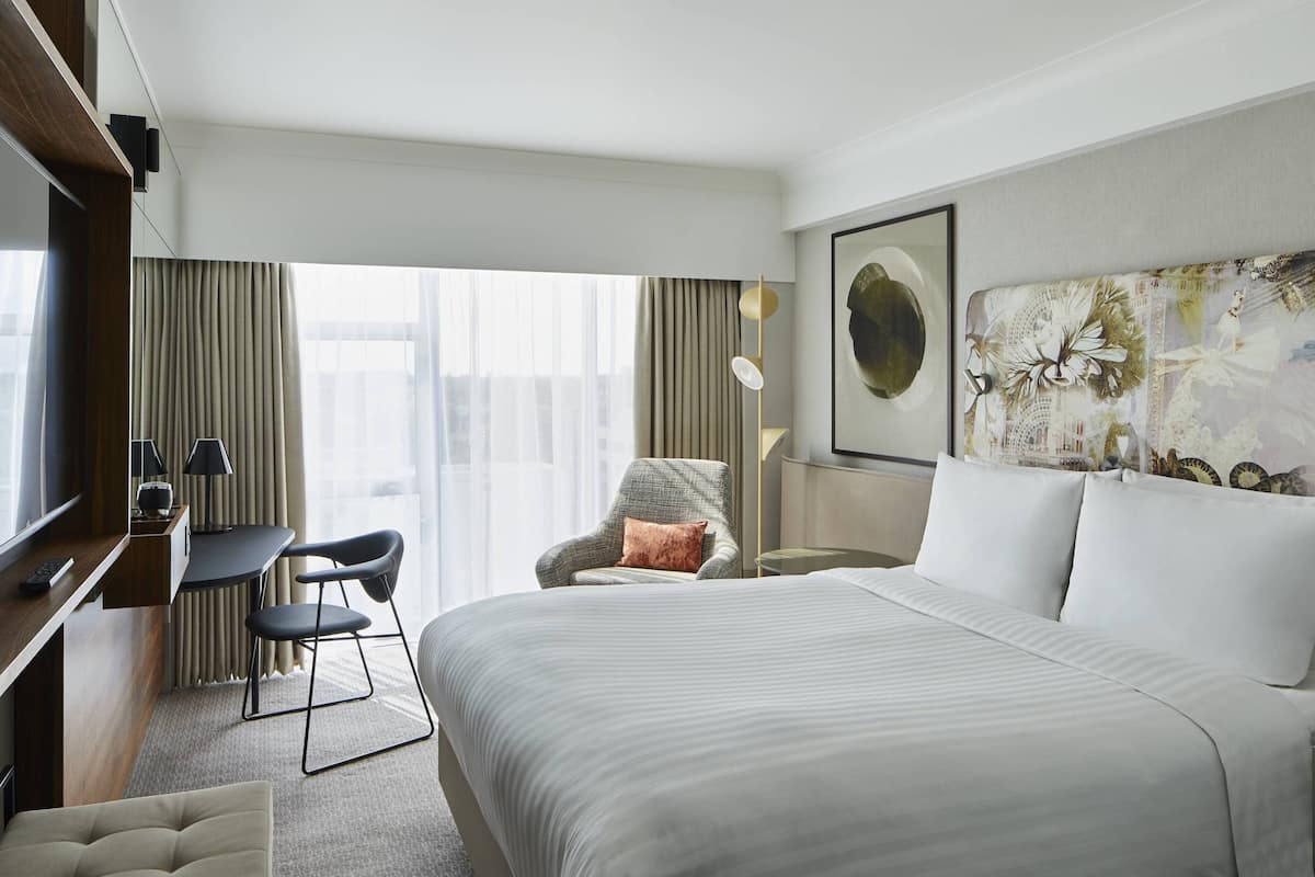 A hotel room with a large bed, a desk with a chair, a small table, and a chair with a cushion.