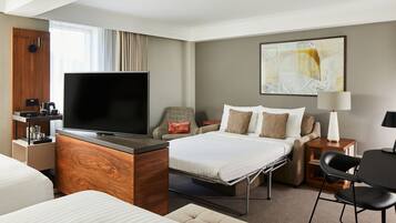 A modern hotel room with a flat-screen TV, a bed with multiple pillows, a desk with a lamp, and a chair.