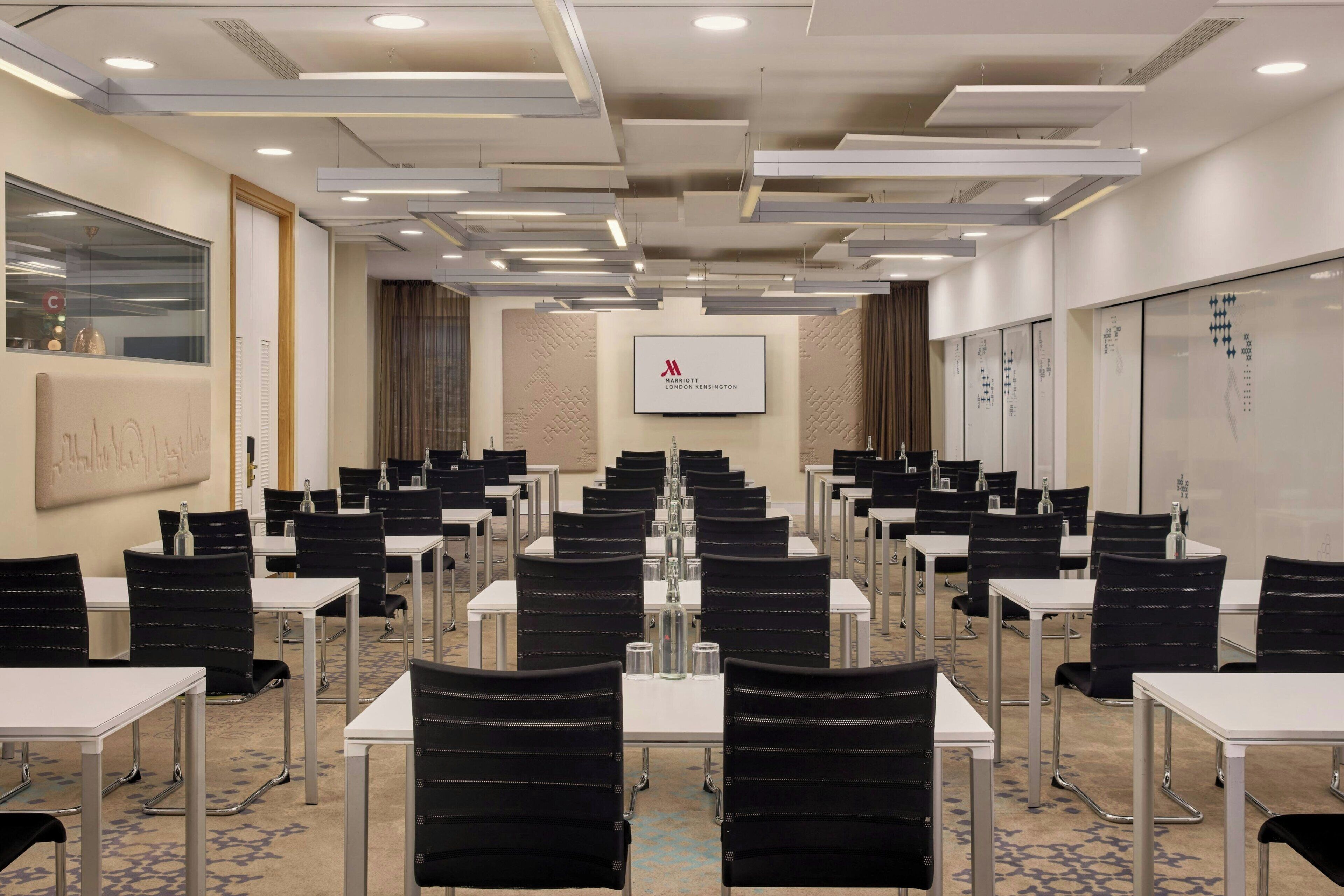 A conference room with rows of tables and chairs, a whiteboard, and a screen displaying a logo.
