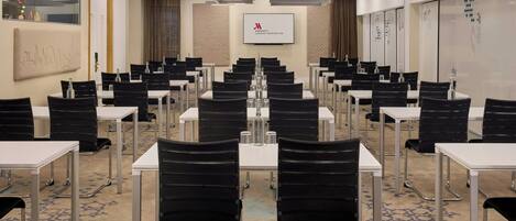 A conference room with rows of tables and chairs, a whiteboard, and a screen displaying a logo.