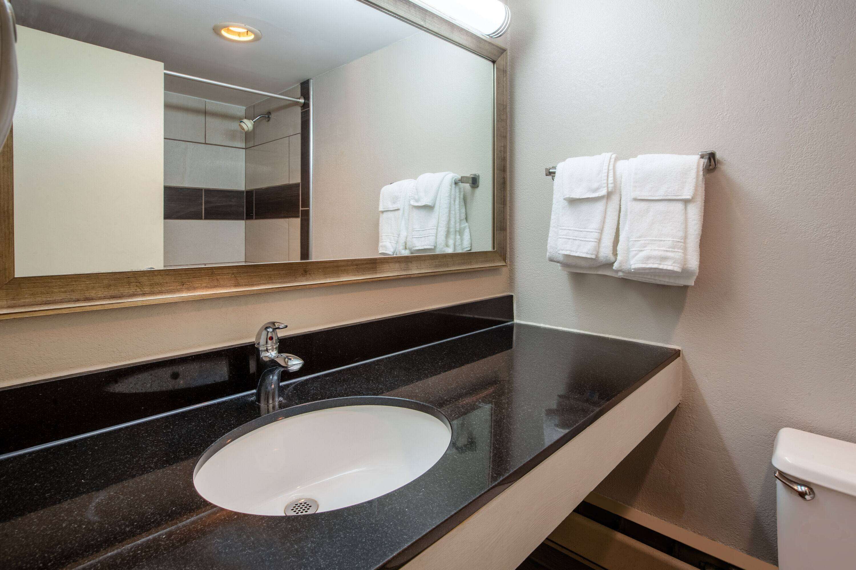 A modern bathroom with a black countertop, a white sink, and a large mirror. There are two white towels hanging on a towel rack.