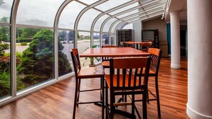 Een modern restaurant met een gebogen glazen dak, houten tafels en stoelen, en uitzicht op de parkeerplaats en de groenvoorziening buiten.
