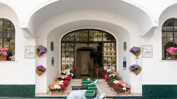 Un edificio blanco con una entrada en arco, una scooter blanca estacionada al frente, y arreglos florales a cada lado.