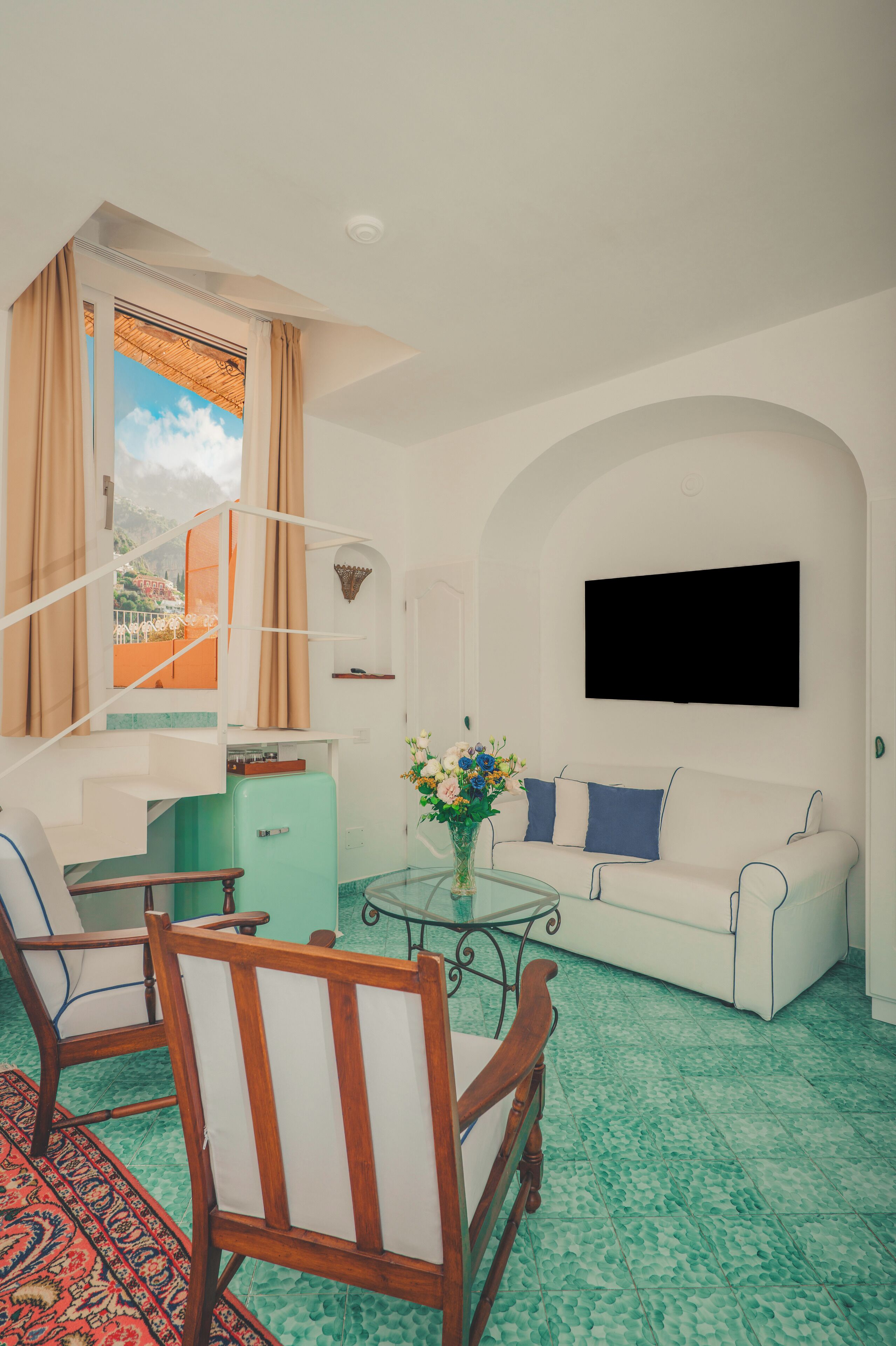 A room with a white sofa, a wooden chair, a small table with a vase, a teal refrigerator, and a window with curtains.