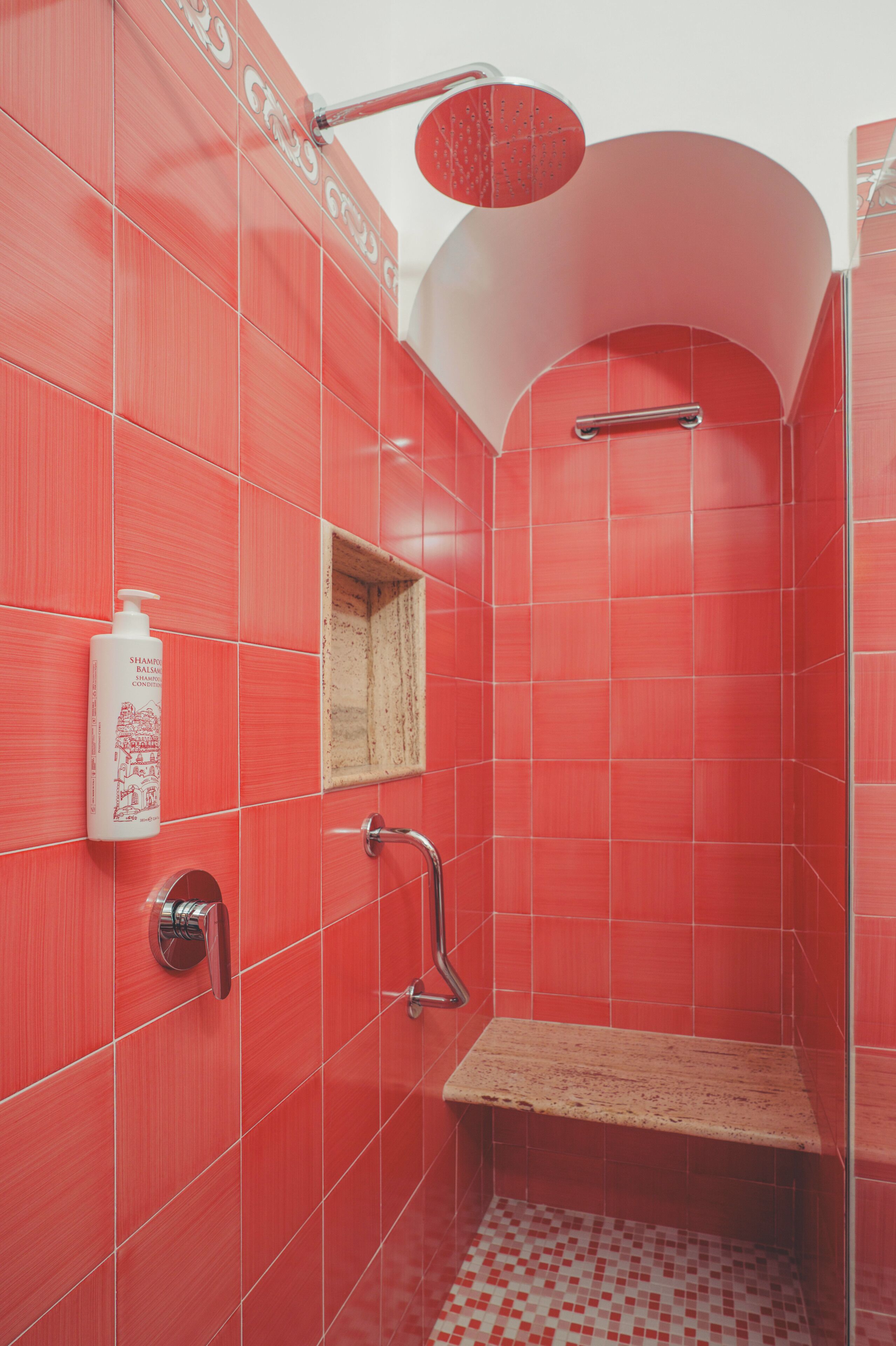 Ducha con azulejos rojos, cabezal de ducha manual, dispensador de jabón y una pequeña estantería de madera.