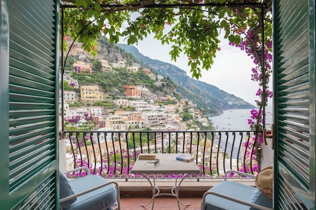 A balcony with a table and chairs, overlooking a coastal town with colorful buildings and a view of the sea.