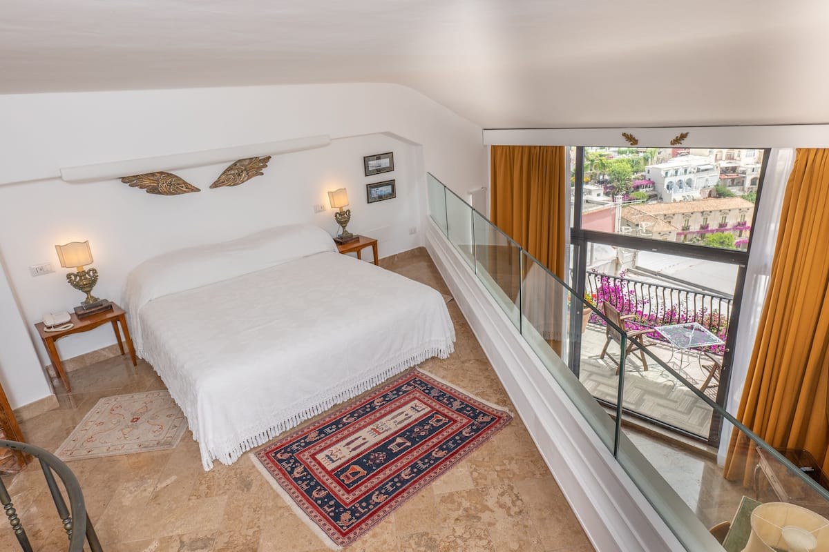 Un dormitorio con cama, lámparas de noche, una alfombra estampada y un balcón con vistas a edificios y flores.