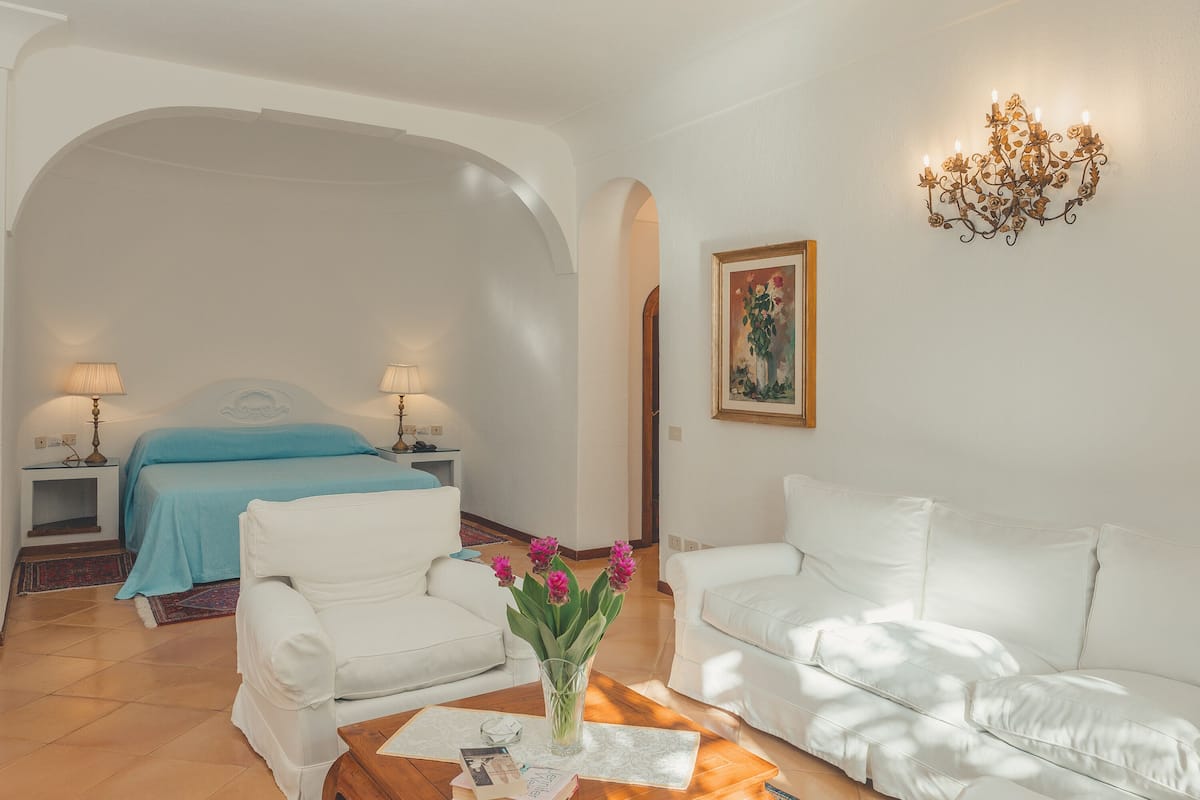 Habitación de hotel con cama, dos sofás blancos, una mesa de centro, una araña y un cuadro en la pared.