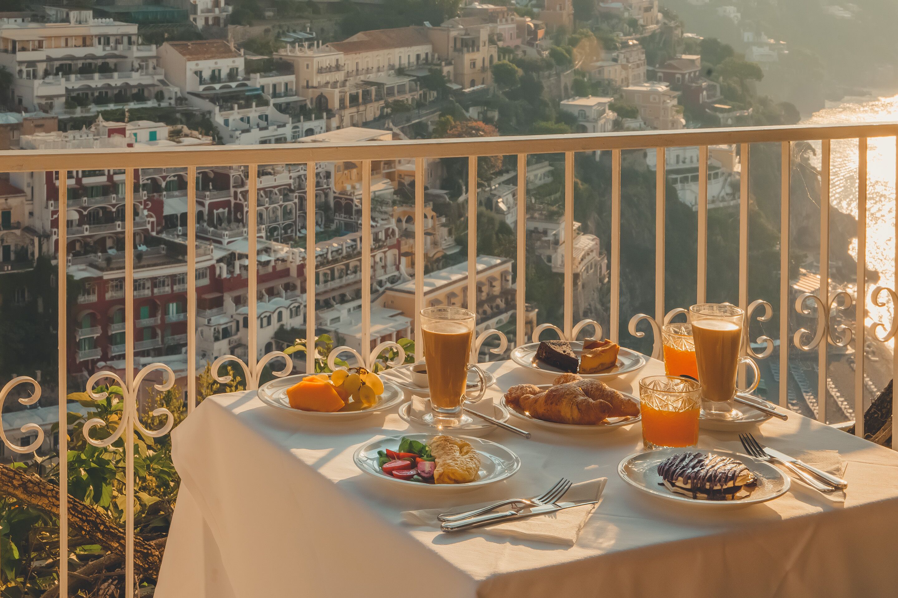 A balcony table with breakfast items and drinks, overlooking a coastal town with white buildings and a view of the sea.