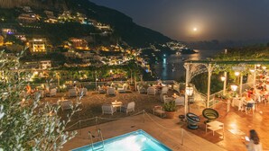 Zona de piscina en la azotea con sillas de descanso y piscina, con vistas a un pueblo costero con edificios y una montaña al fondo.