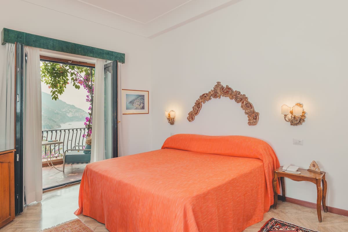 Un dormitorio con una colcha naranja, una mesita de noche de madera, un cuadro enmarcado en la pared y un balcón con vistas a las montañas.