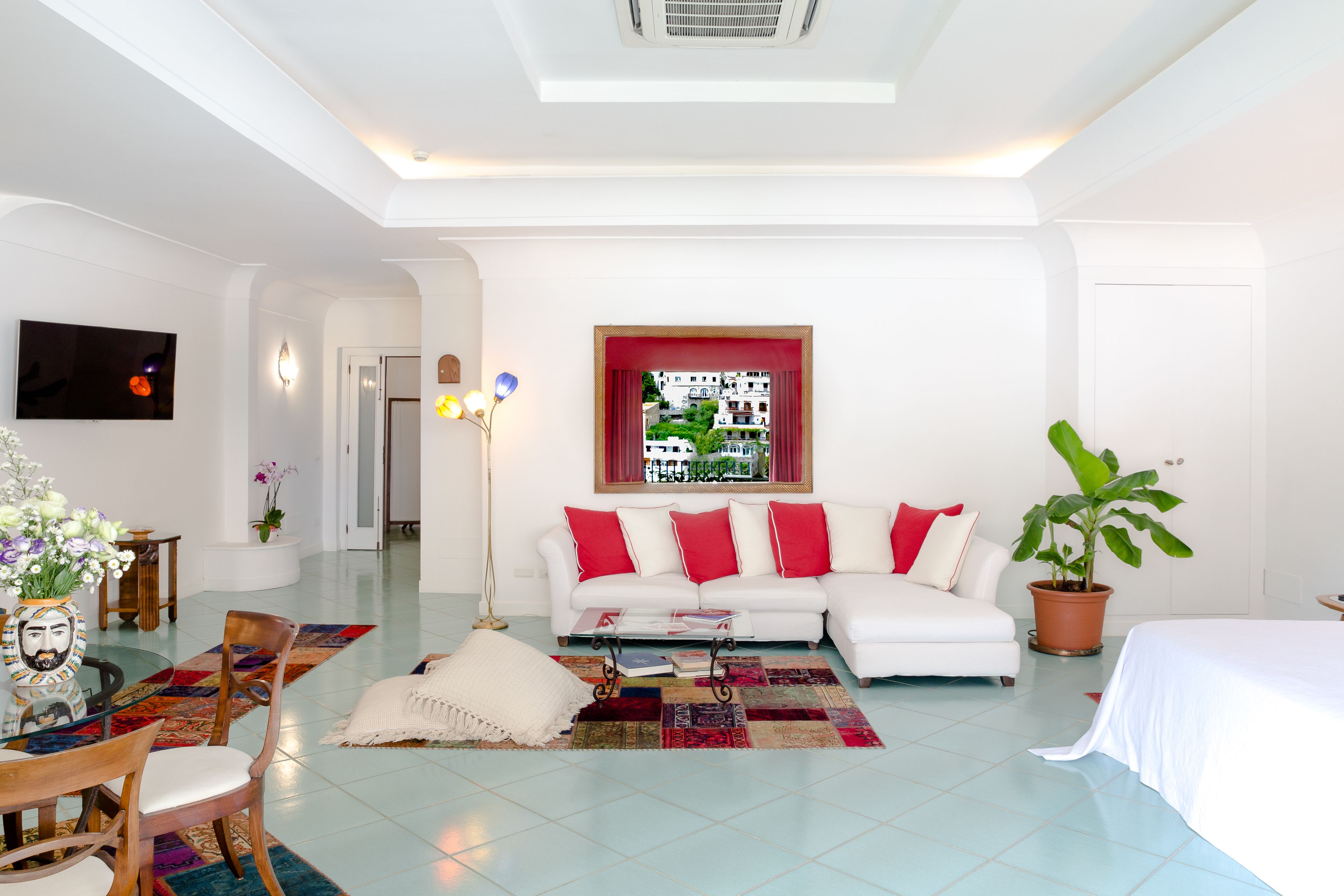 Un salon moderne avec un canapé blanc, des coussins rouges, une table basse avec des magazines, un téléviseur fixé au mur, une grande fenêtre donnant sur des immeubles et une plante en pot.