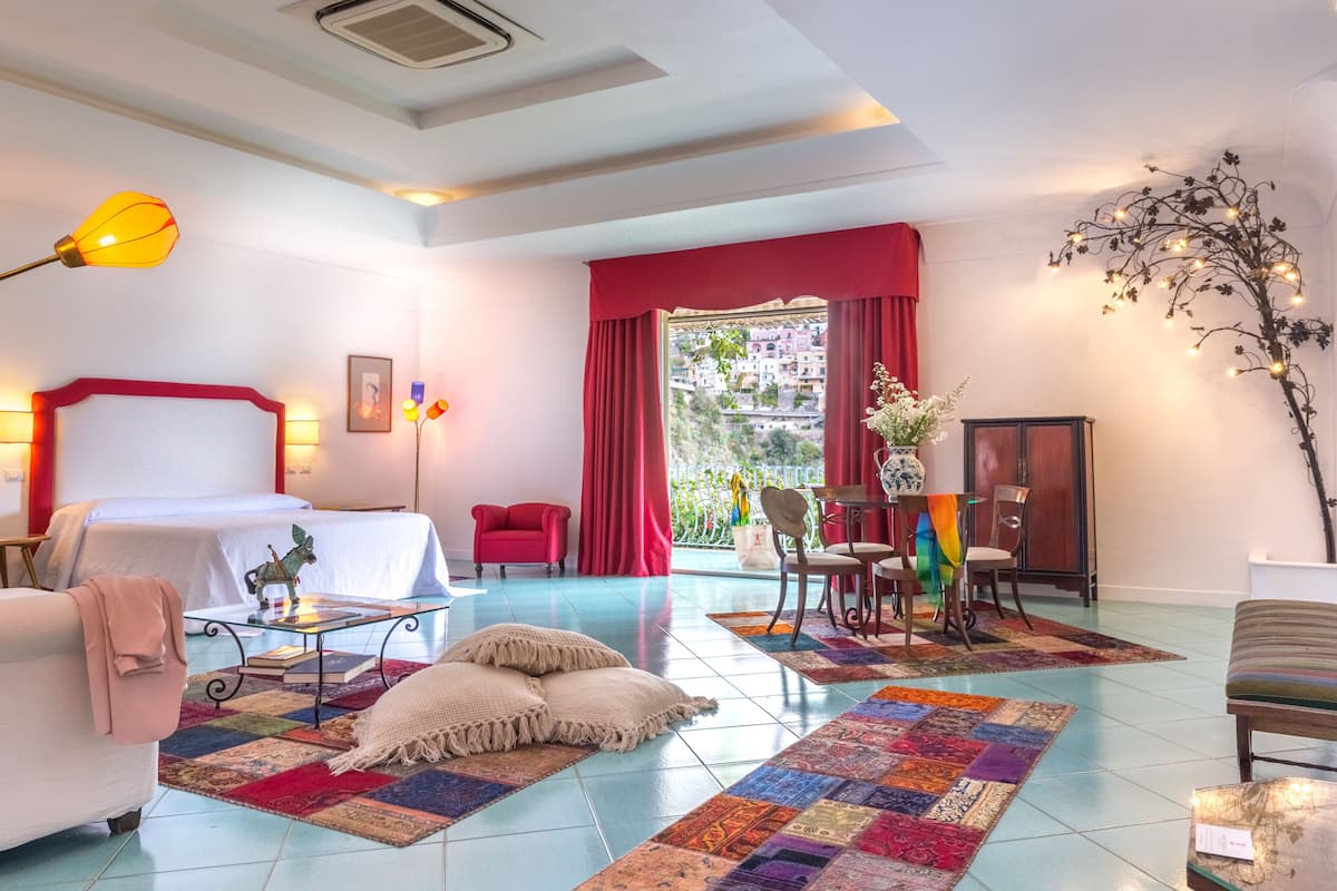 Habitación de hotel con cama, una silla roja, una mesa con sillas, una mesita con un florero y vistas al exterior.
