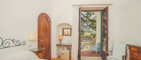 Quarto com cama, escrivaninha, cadeira e guarda-roupa. Há uma porta que dá para área externa com vista para árvores e uma cadeira azul.
