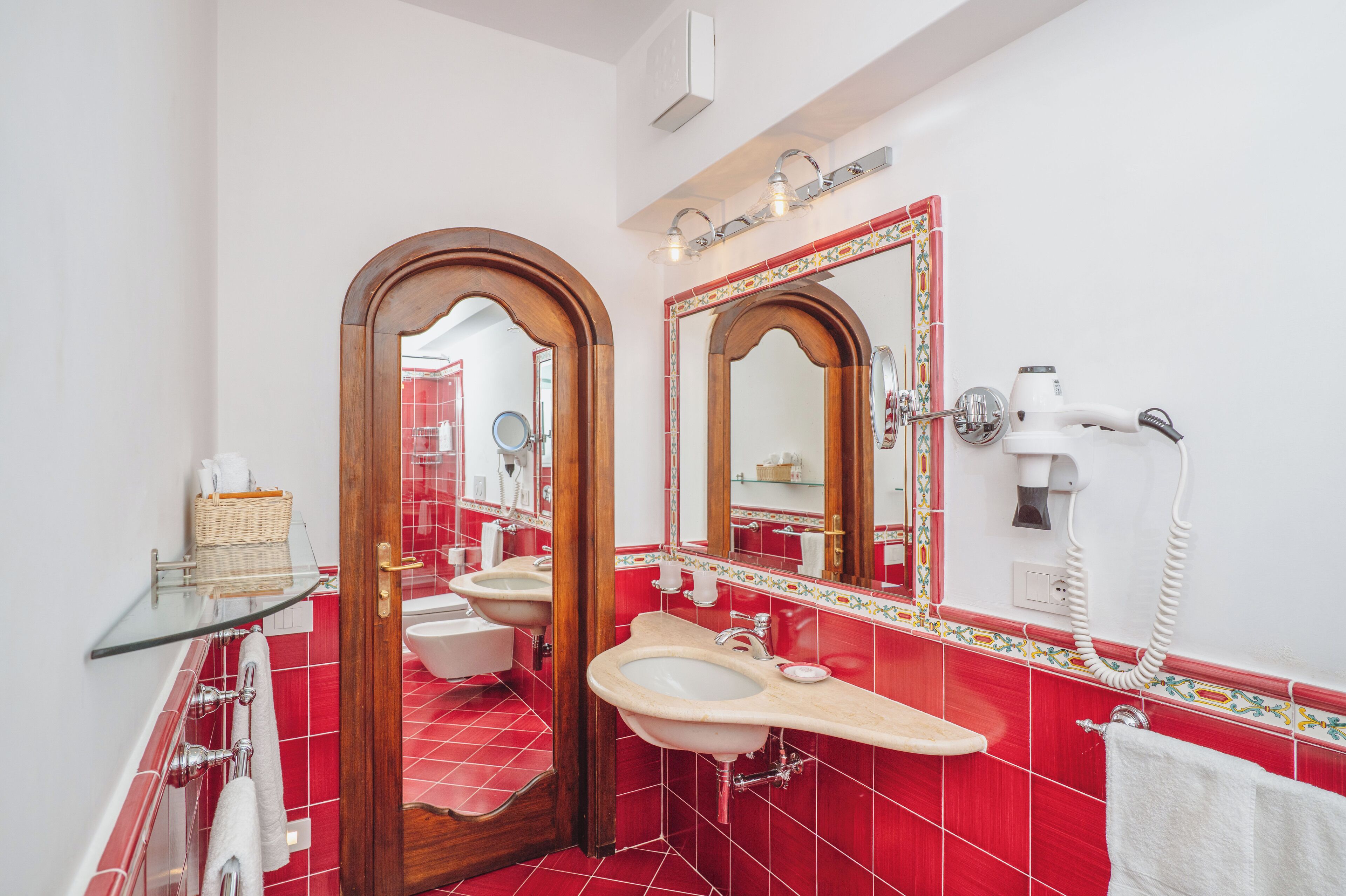 A bathroom with a red tiled floor, a wooden door, a white sink, a mirror, and a hairdryer.