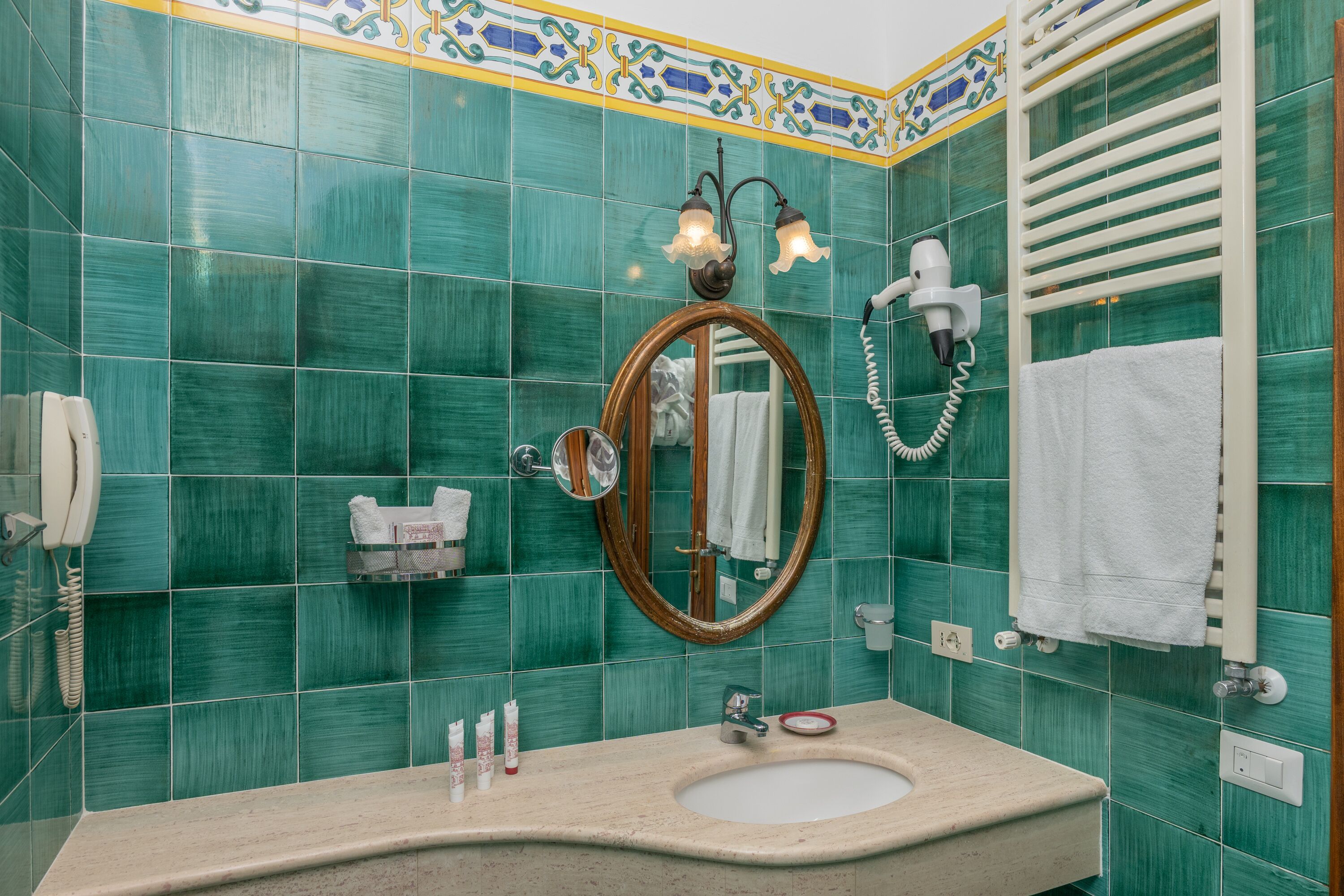 A bathroom with green tiles, a sink, a mirror, a hairdryer, and a towel rack.