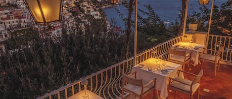 Terraza con mesas y sillas dispuestas para comer, con vistas a un pueblo costero y al mar.