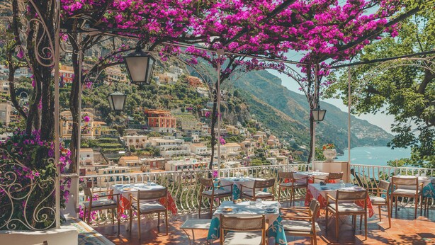 Una terraza con mesas y sillas, con vistas a un pueblo costero con edificios y montañas.