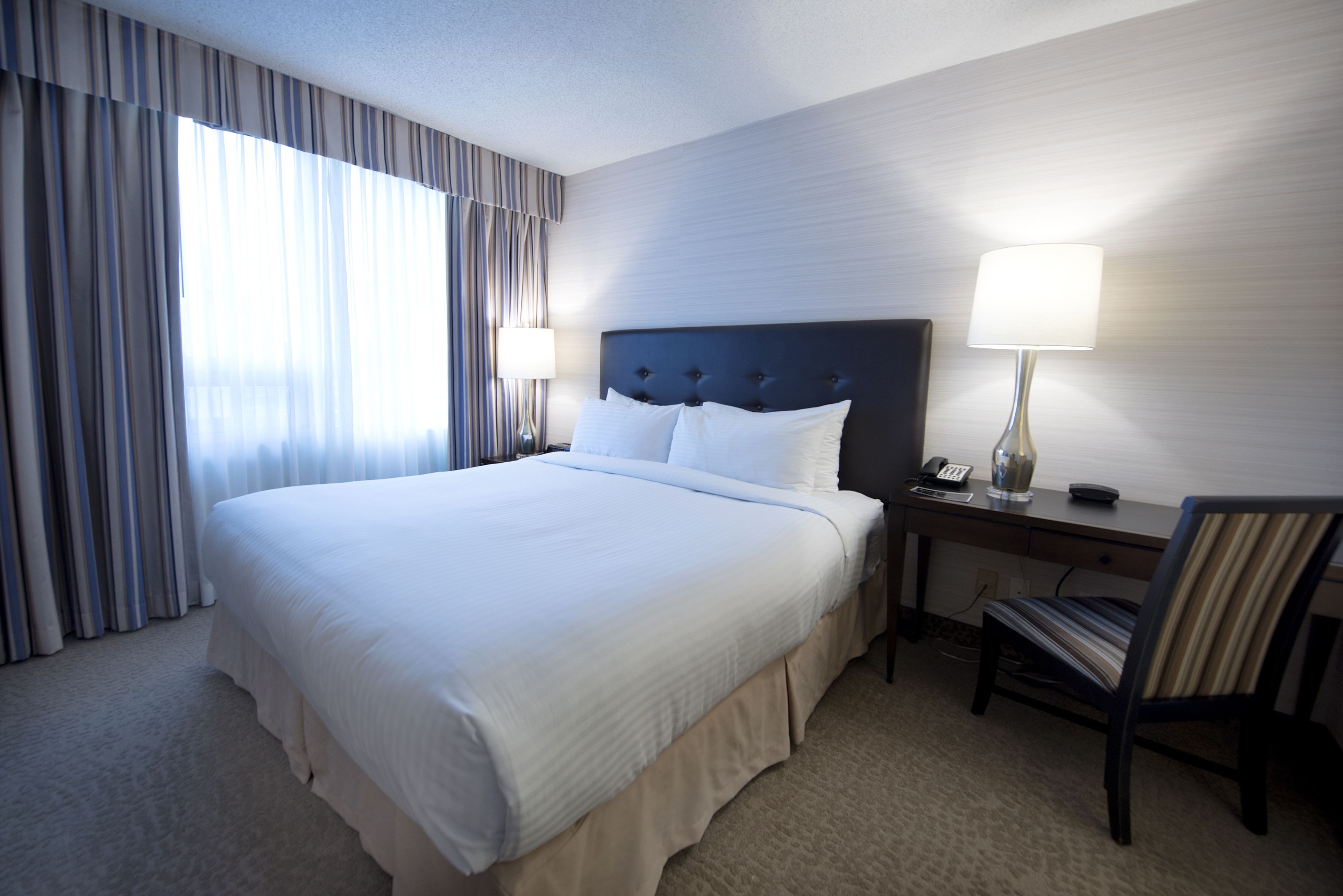 Quarto de hotel com uma cama grande, uma escrivaninha com abajur, uma cadeira e uma janela com cortinas.