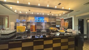 Zona de bufé con varias estaciones de comida, incluyendo un bol de fruta y un dispensador de zumo.