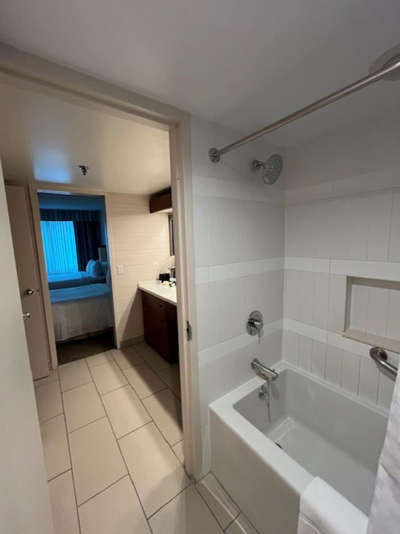 Et badeværelse med badekar, bruser og toilet. Der er en dør til et soveværelse med seng og et vindue med gardiner.