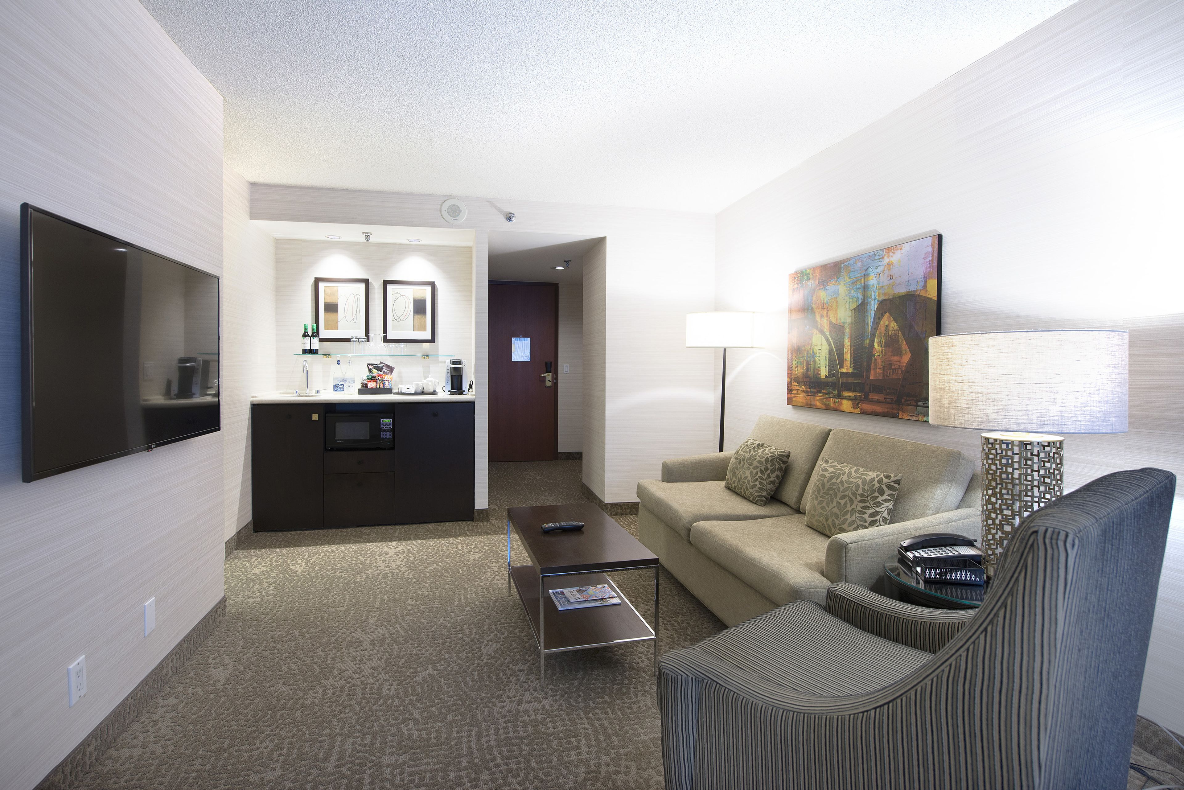 A hotel room with a flat-screen TV, a sofa, a coffee table, a lamp, and a painting on the wall.
