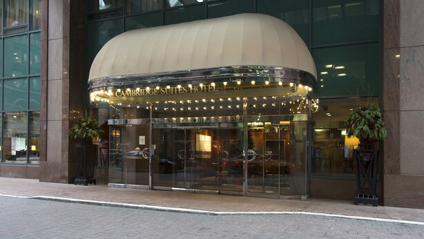 Entrada del Hotel Cambridge Suites con fachada de vidrio, un toldo curvo y el reflejo de coches en el cristal.