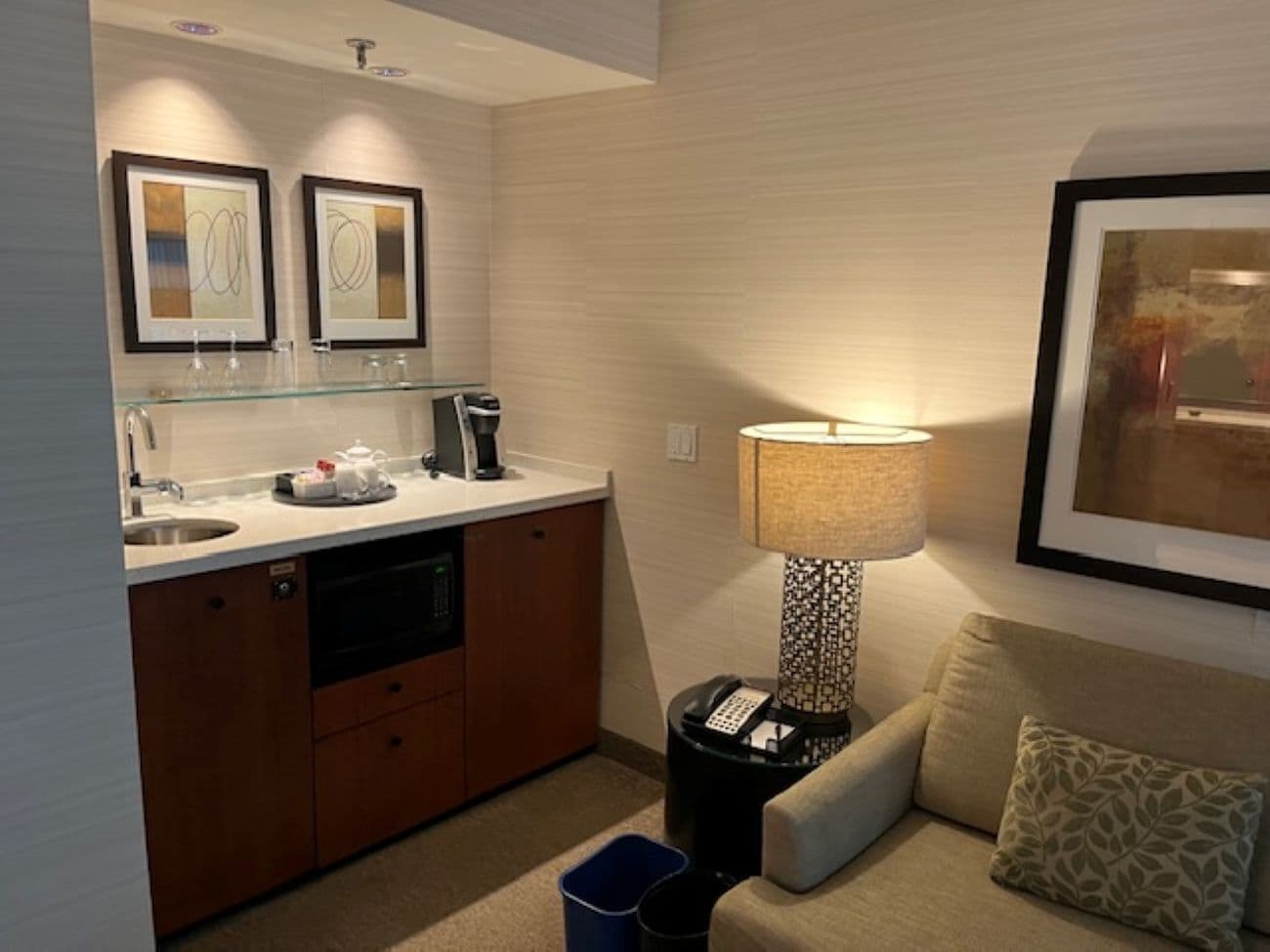 Habitación de hotel compacta con kitchenette, sofá, lámpara y cuadros en la pared.