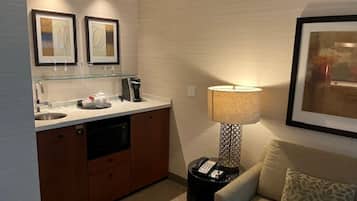 Habitación de hotel compacta con kitchenette, sofá, lámpara y cuadros en la pared.