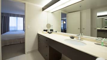 A hotel bathroom with a large mirror, a sink, and a bed in the adjacent room.