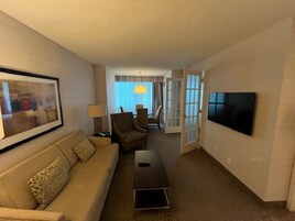Een hotelkamer met een beige bank, een salontafel, een aan de muur bevestigde televisie en een deur naar een balkon.