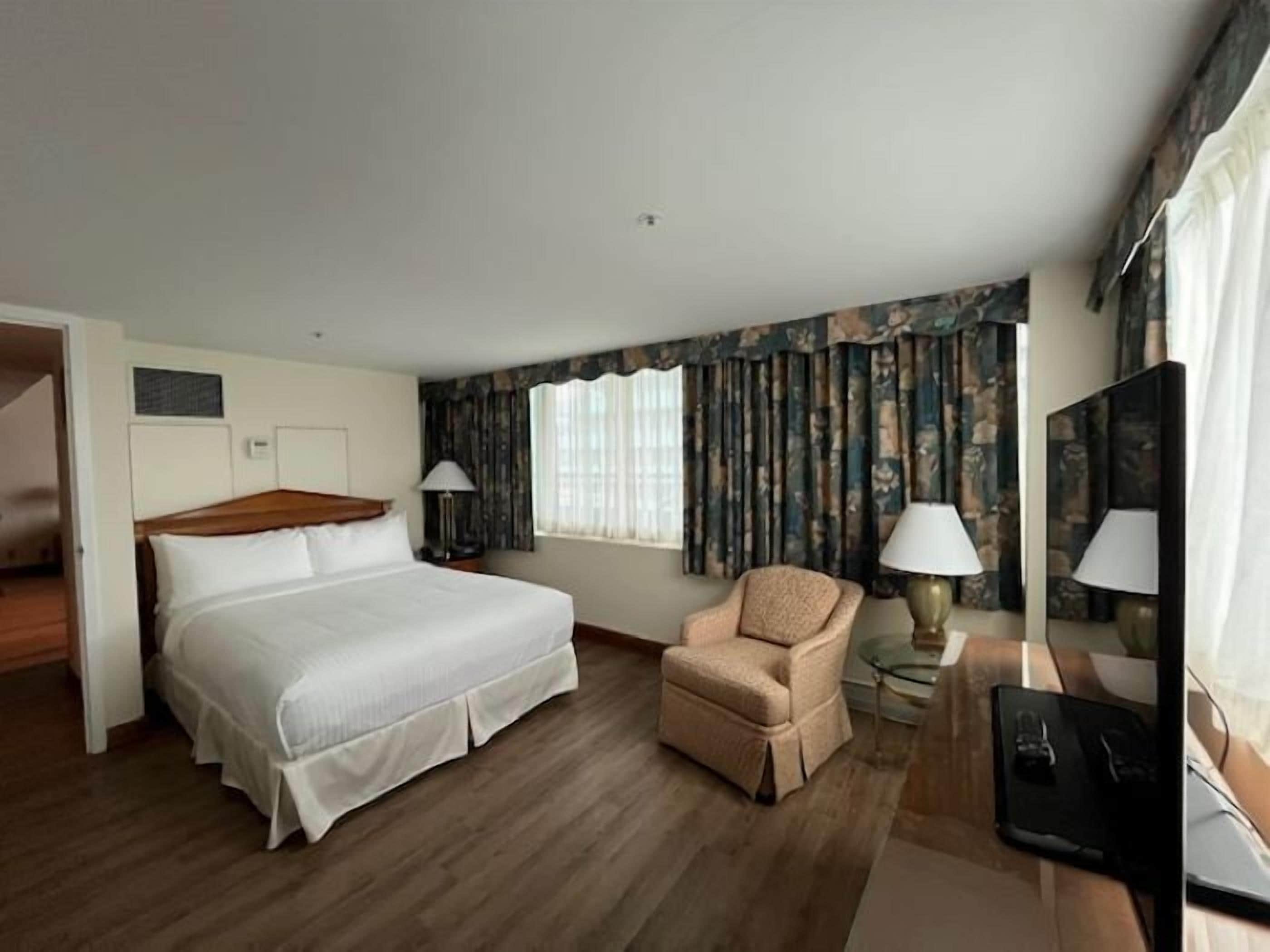 A hotel room with a large bed, a chair, a desk with a computer, and a window with curtains.