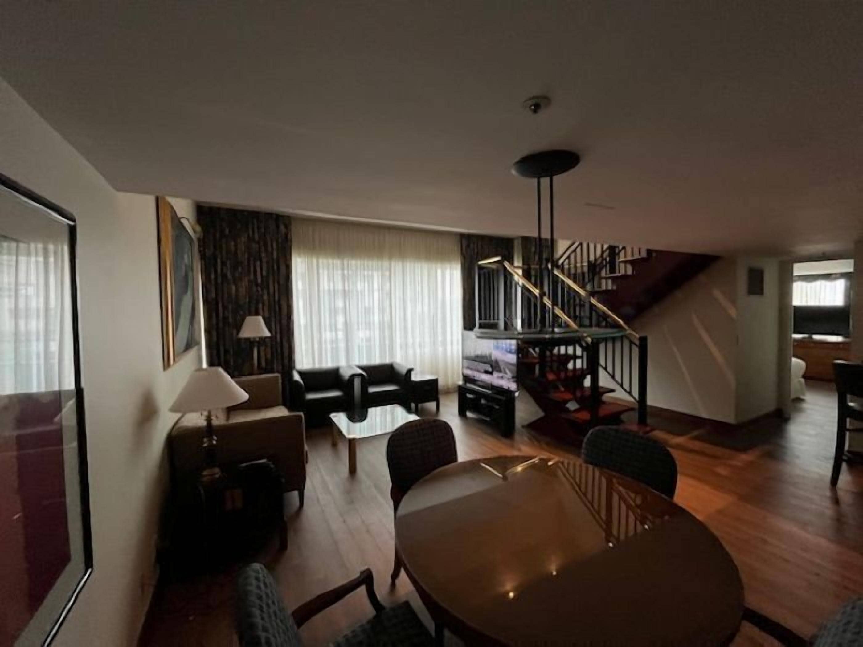 A spacious living area with a round wooden table, two sofas, and a coffee table. A staircase with a railing is visible in the background.