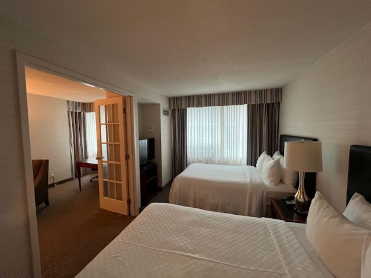 A hotel room with two beds, a desk, a chair, a television, and a door leading to another room.