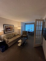 Habitación de hotel con sofá, sillón, mesa de centro y un área de comedor con mesa y sillas.