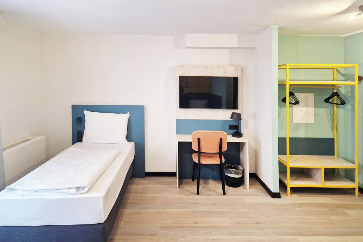 Habitación de hotel con cama, escritorio con silla, televisor montado en la pared y un mueble de almacenamiento amarillo.