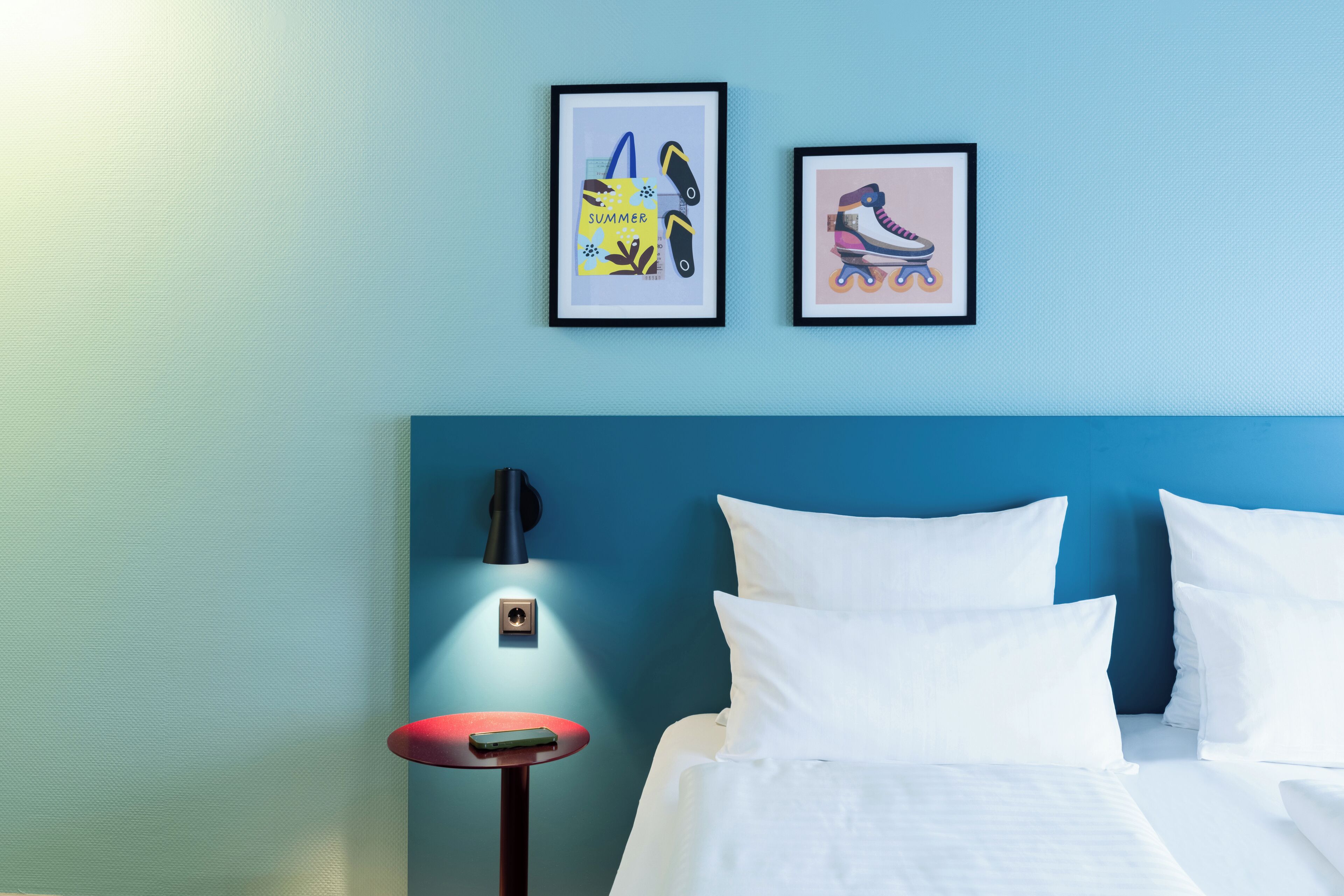 A hotel room with a blue wall, a bed with white linens, a red side table, and two framed pictures on the wall.
