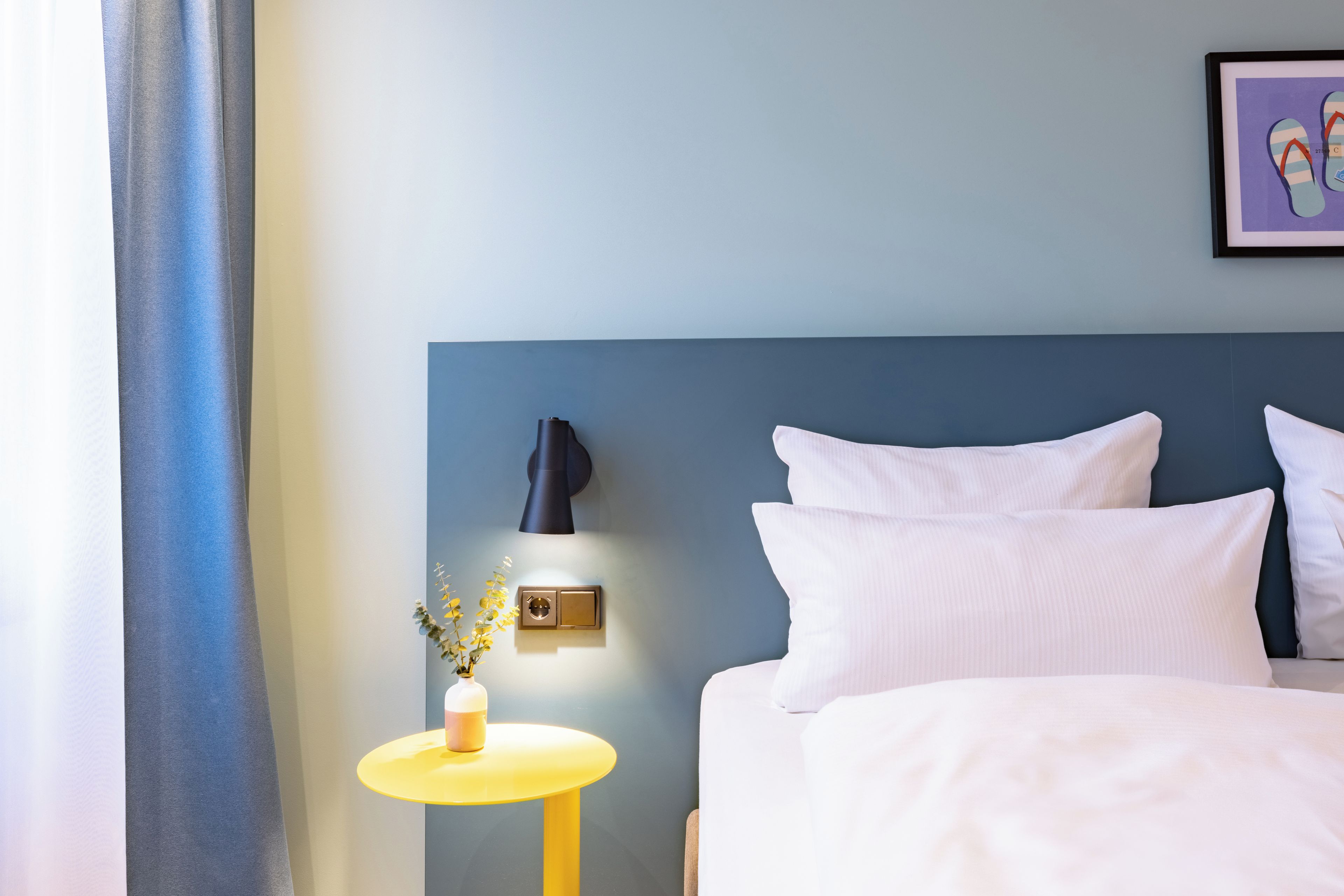 Habitación de hotel con cama, mesita de noche con lámpara, luz empotrada en la pared y un cuadro enmarcado.