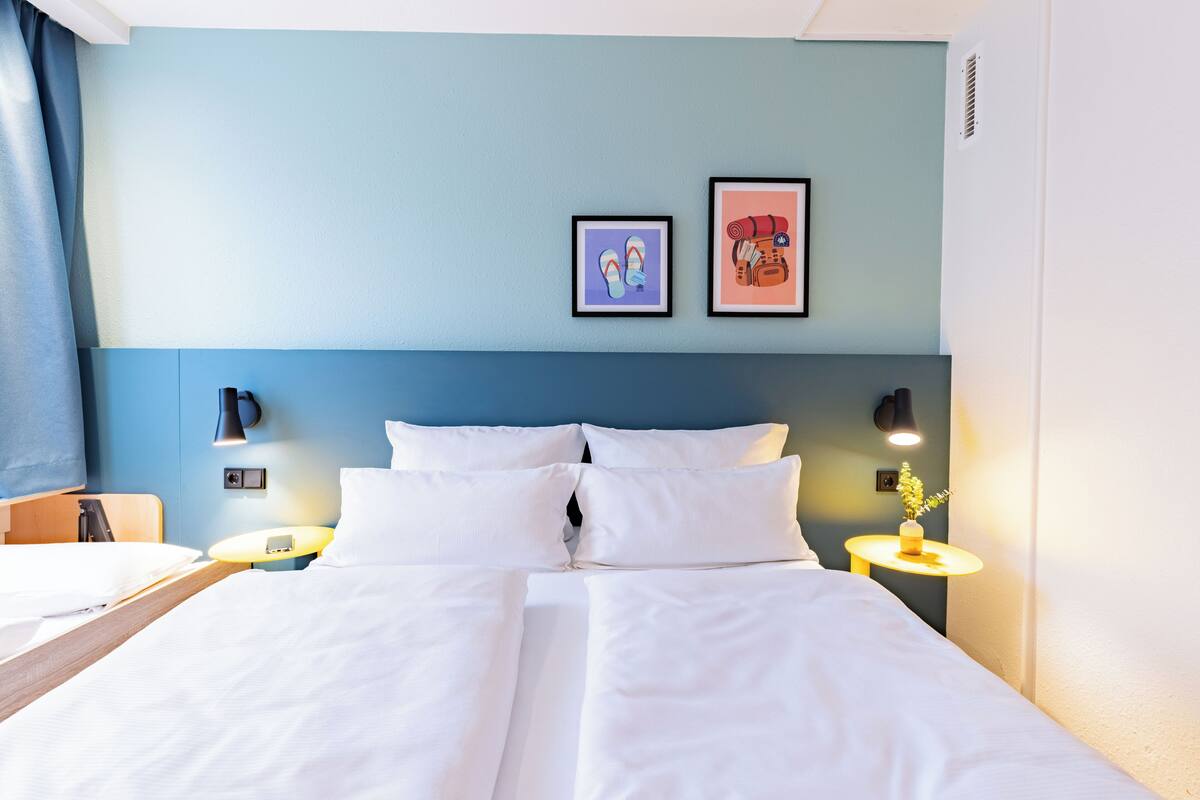 Habitación de hotel con una cama, dos mesitas de noche, una lámpara y dos cuadros en la pared.