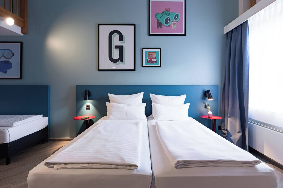 Una habitación de hotel con dos camas individuales, una letra 'G' grande enmarco y una pequeña foto enmarco de un robot.