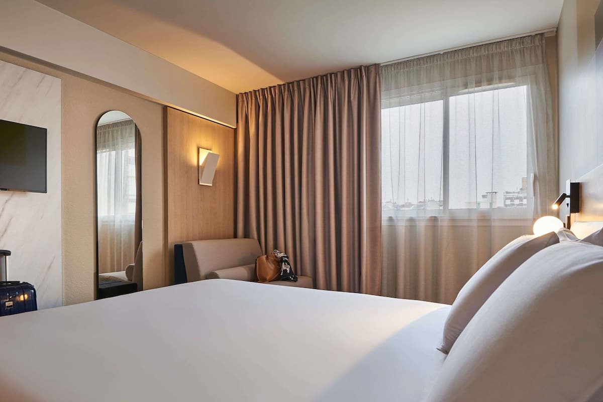 A hotel room with a large bed, a flat-screen TV, a wall-mounted mirror, a bedside table with a lamp, and a view of the city through the window.