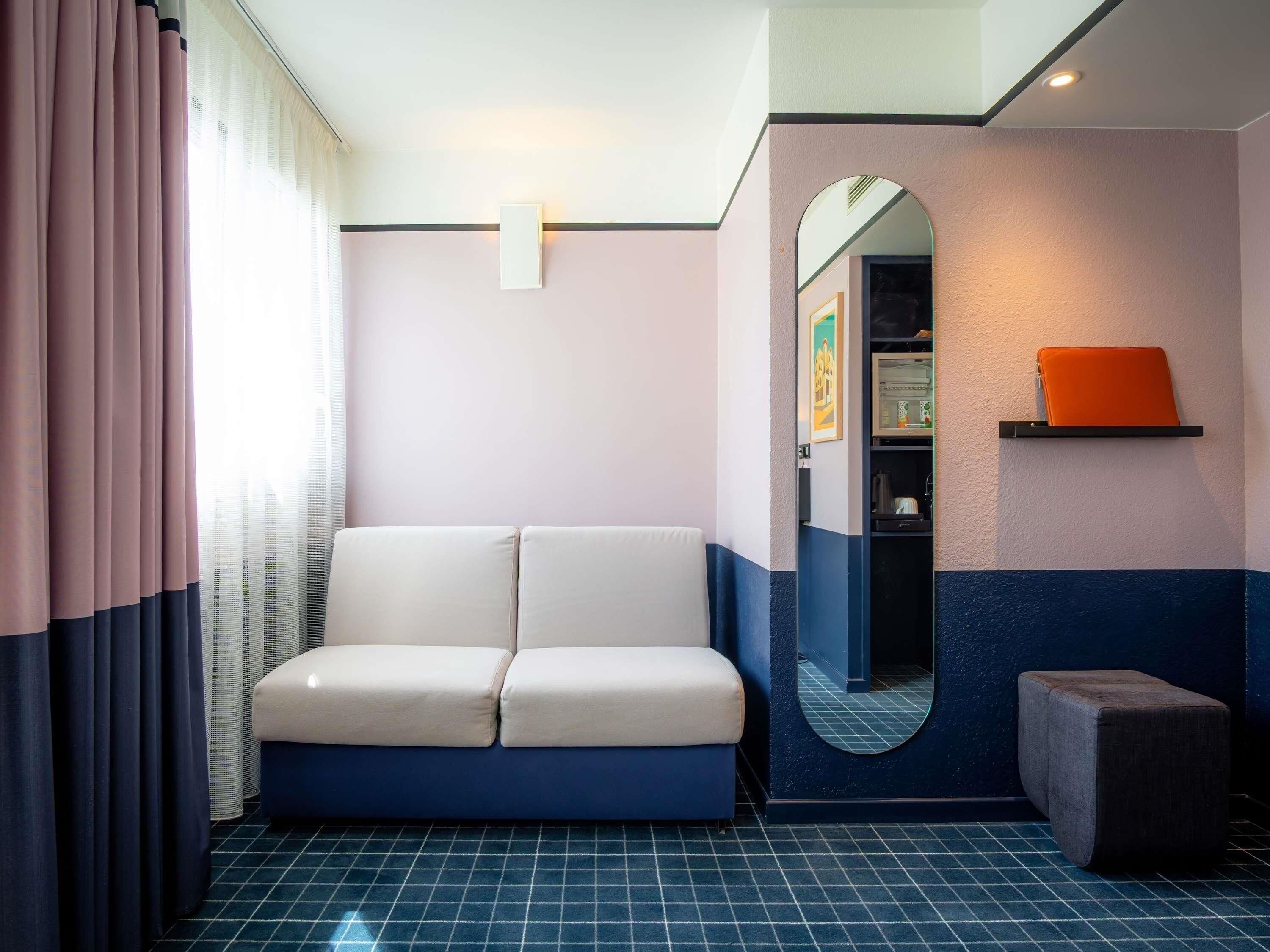 Ein Zimmer mit einer Couch, einem kleinen Hocker, einem Spiegel und einem Regal mit einem Buch.