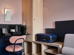 Habitación pequeña con escritorio, silla, cafetera, maleta y cama.