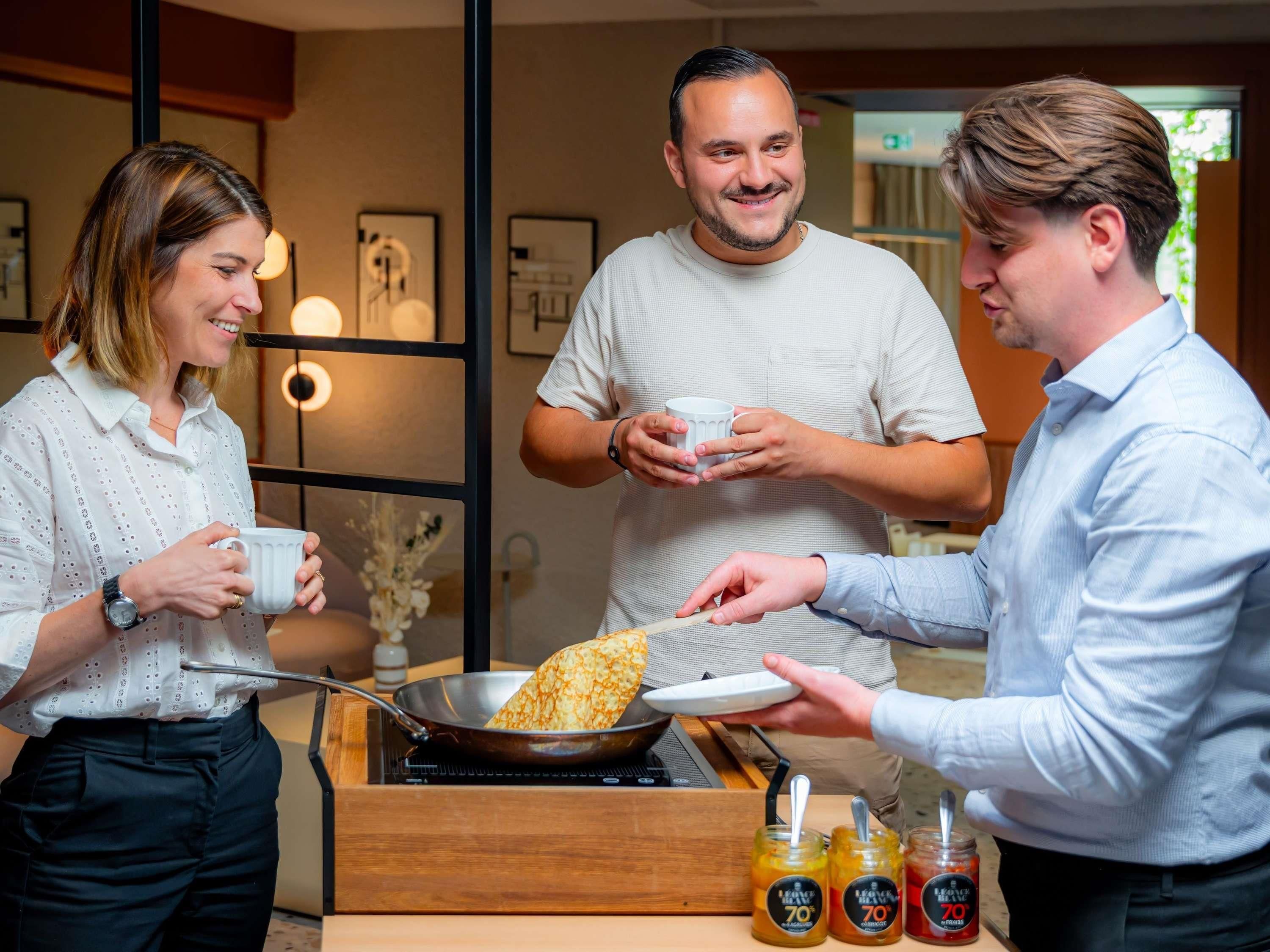 Uma pessoa servindo uma crepe em uma frigideira para duas pessoas segurando xícaras.